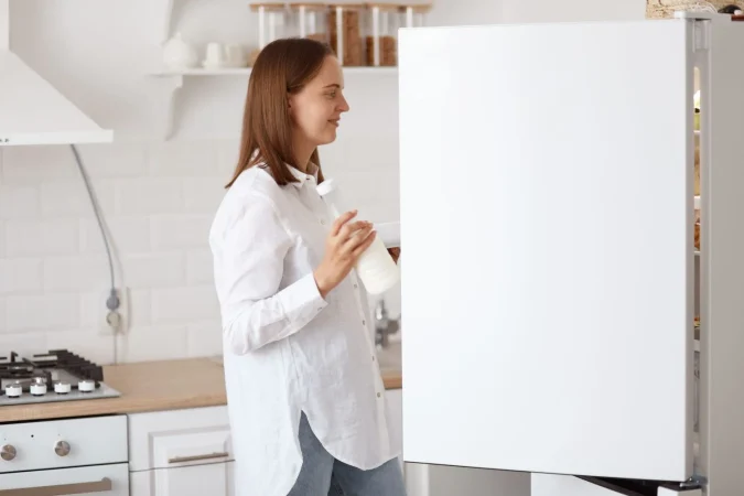profile portrait attractive dark haired woman wearing white shirt looking smiling inside fridge with positive emotions holding plate hands posing with kitchen set background Merca2.es