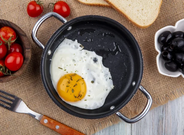 cerrar vista de huevo frito en sarten con tenedor y tazon de rodajas de tomate y pan con aceitunas sobre tela de saco y superficie de madera Merca2.es