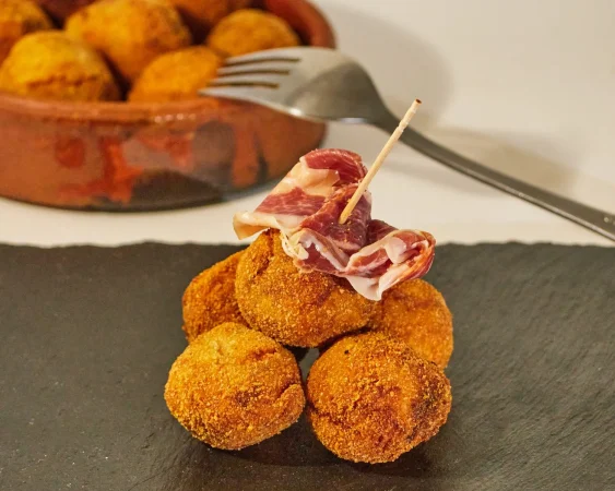 pincho de varias croquetas redondas con una rebanada de jamon iberico en una tabla de pizarra negra Merca2.es