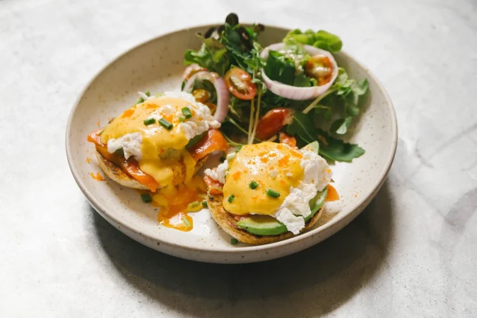Huevos benedictinos, el arte del desayuno perfecto que nunca pasa de moda 2 Merca2.es huevo benedict con salmon y aguacate servido con ensalada en plato blanco Merca2.es
