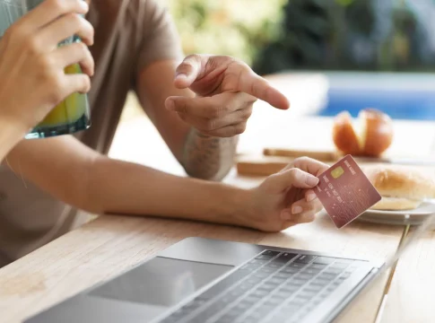 compras-online-laptop-tarjeta-credito Primer plano de una persona sosteniendo una tarjeta de crédito frente a una laptop mientras otra señala la pantalla durante una compra online.