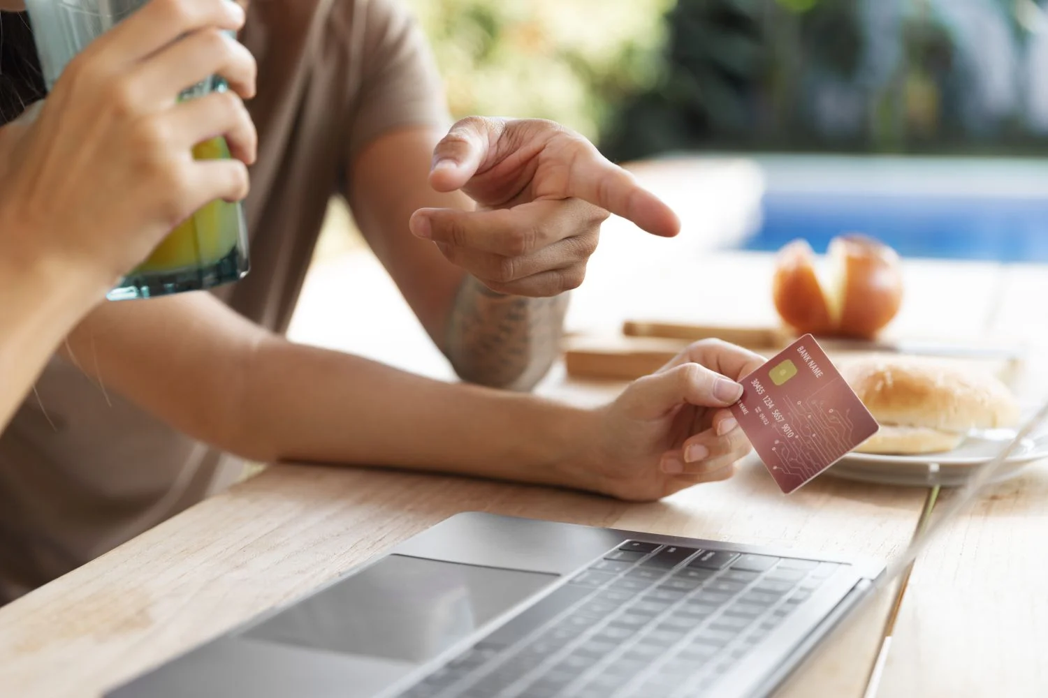 compras-online-laptop-tarjeta-credito Primer plano de una persona sosteniendo una tarjeta de crédito frente a una laptop mientras otra señala la pantalla durante una compra online.