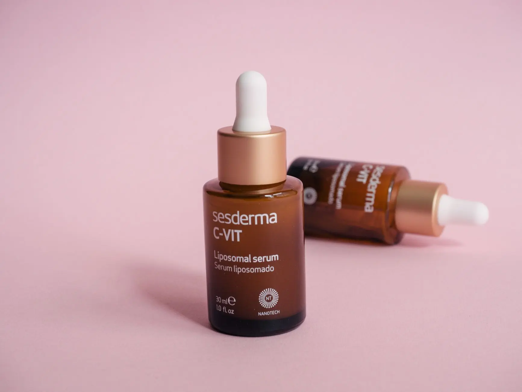 Primer plano de dos botellas de suero cosmético con gotero sobre una superficie rosa.