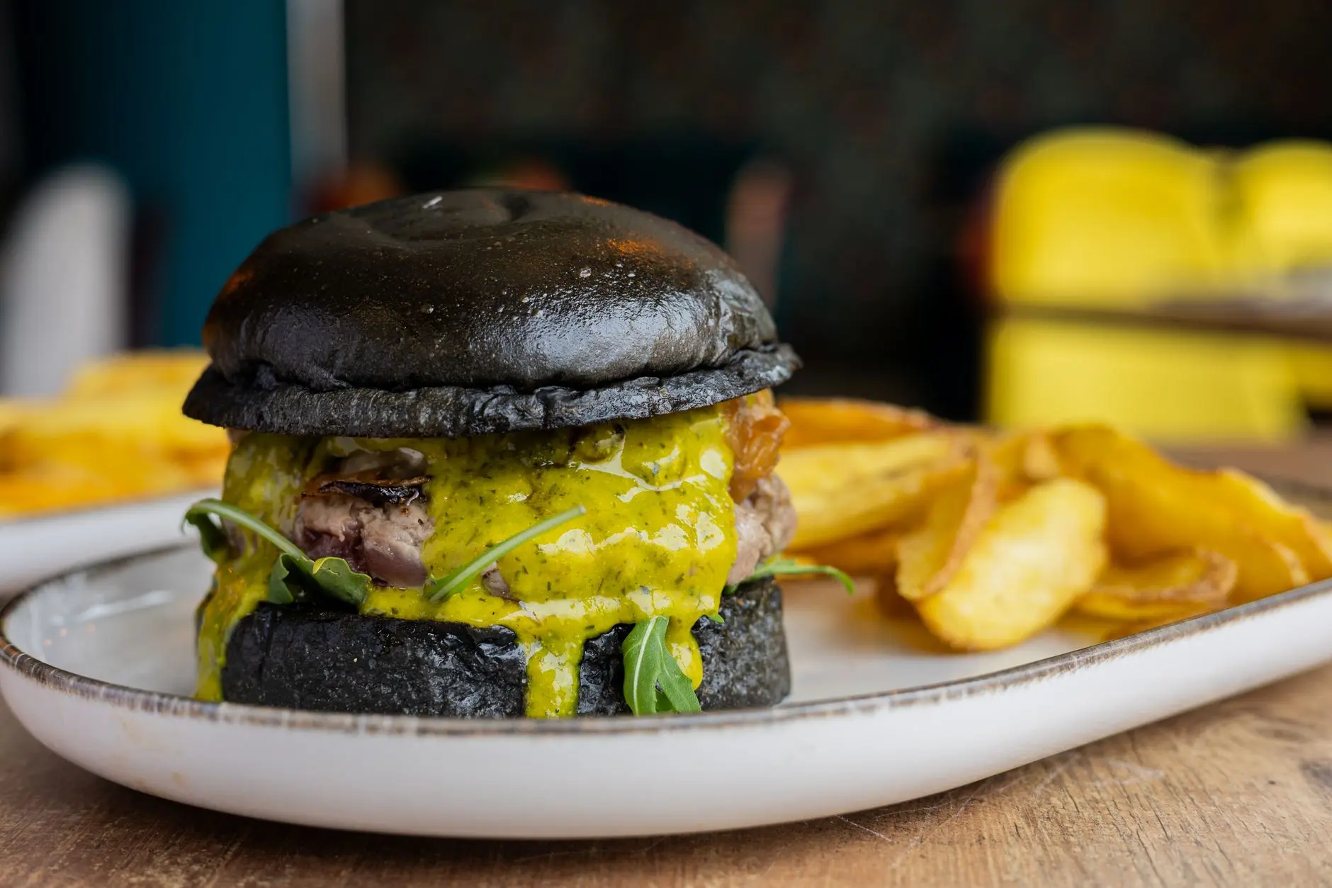 Primer plano de una hamburguesa gourmet con pan negro y salsa de mostaza servida con patatas fritas en un plato de cerámica.