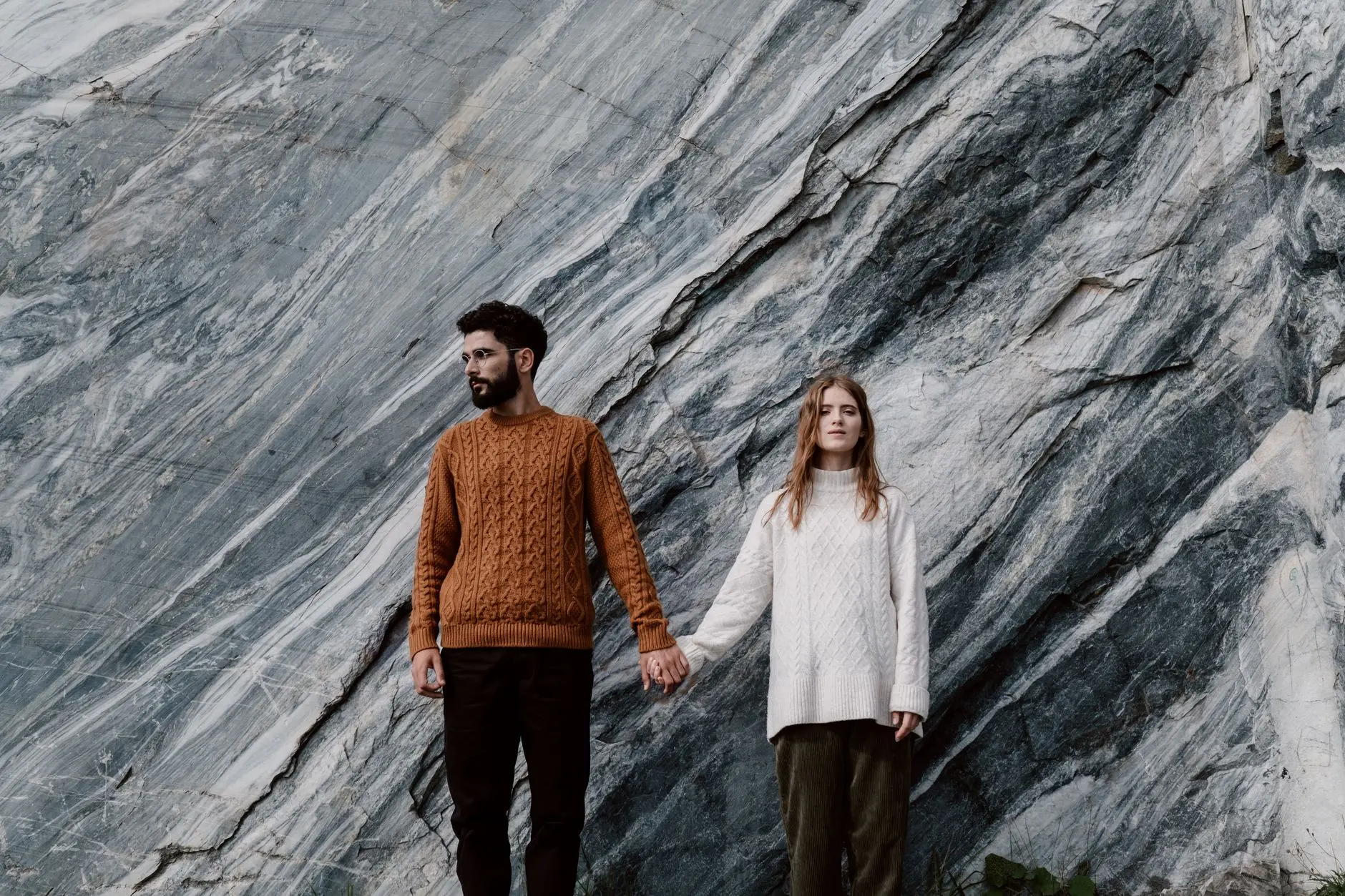 Una pareja en suéteres de punto, de pie junto a una formación rocosa de mármol, tomados de la mano.