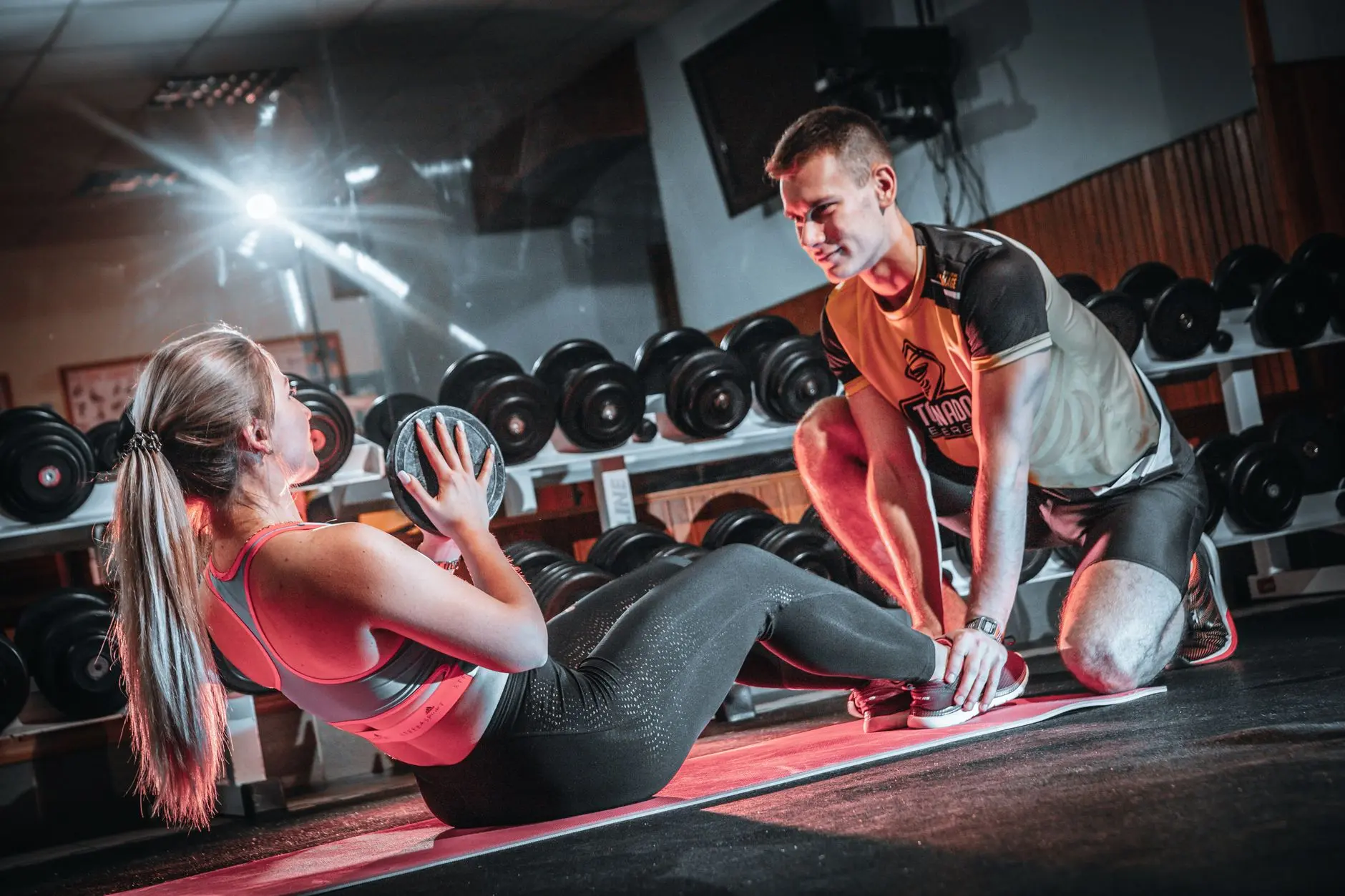 Un entrenador personal asiste a una mujer que hace abdominales con una pelota medicinal en un entorno de gimnasio.