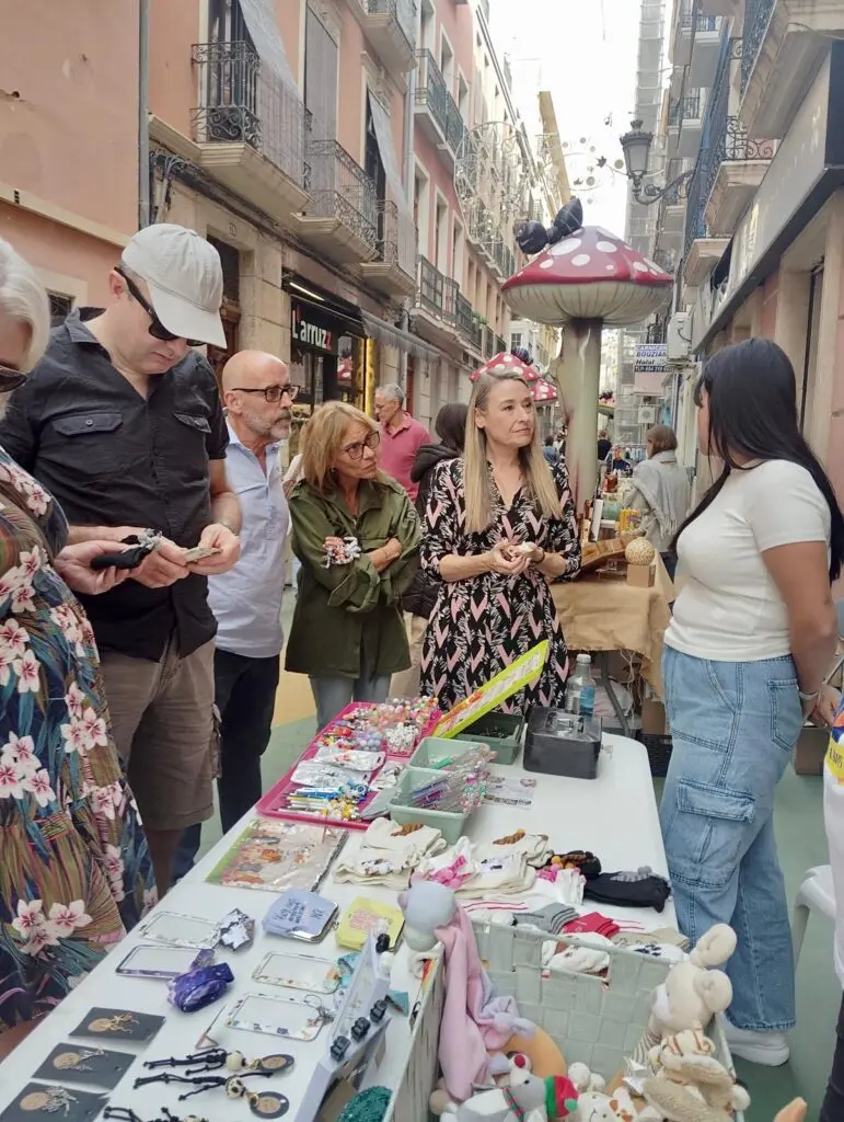 Puesto callejero en Botigues al Carrer Varias personas miran accesorios y artículos variados en un puesto callejero bajo adornos de setas.