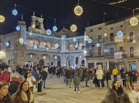 Plaza iluminada con decoración navideña Plaza con edificios de piedra, luces navideñas colgantes y numerosas personas.