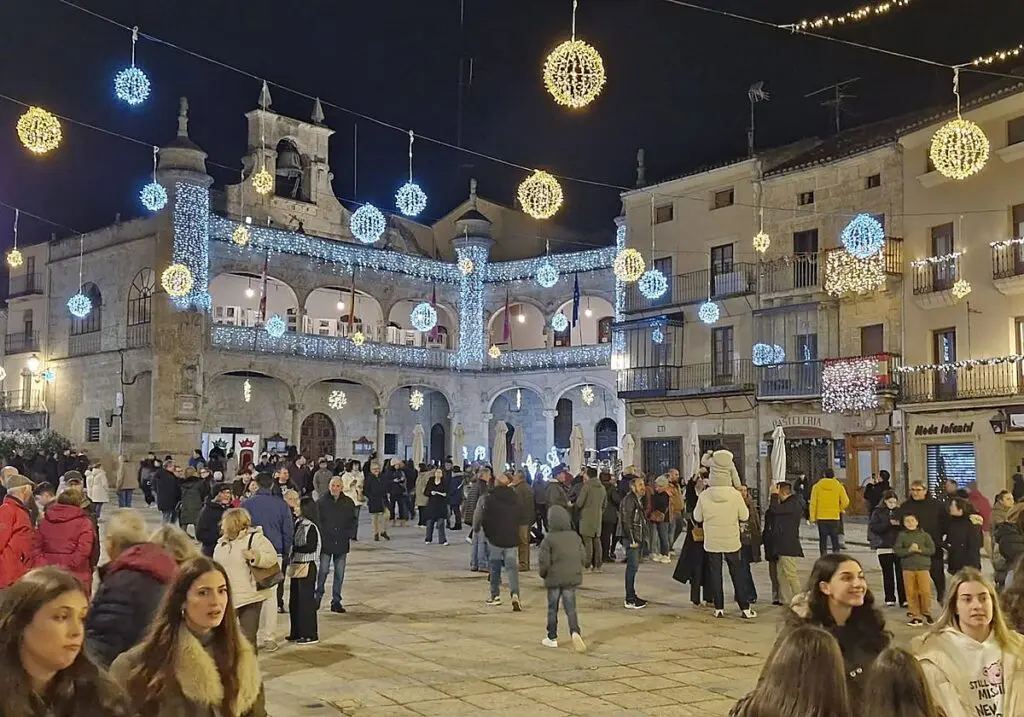 Plaza iluminada con decoración navideña Plaza con edificios de piedra, luces navideñas colgantes y numerosas personas.