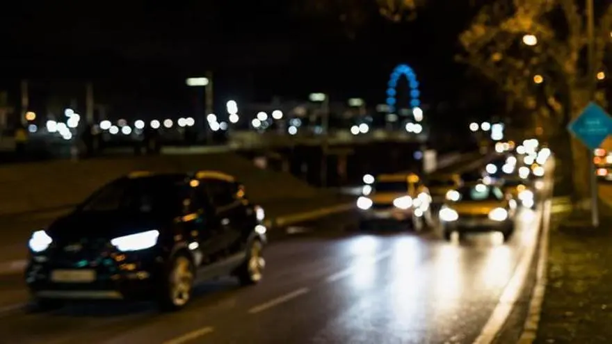 Varios coches circulan de noche por una vía urbana con los faros encendidos.