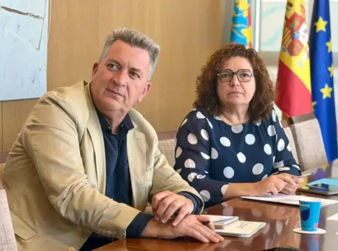 Dos personas en una mesa con documentos Dos personas sentadas a una mesa, con documentos y banderas al fondo.
