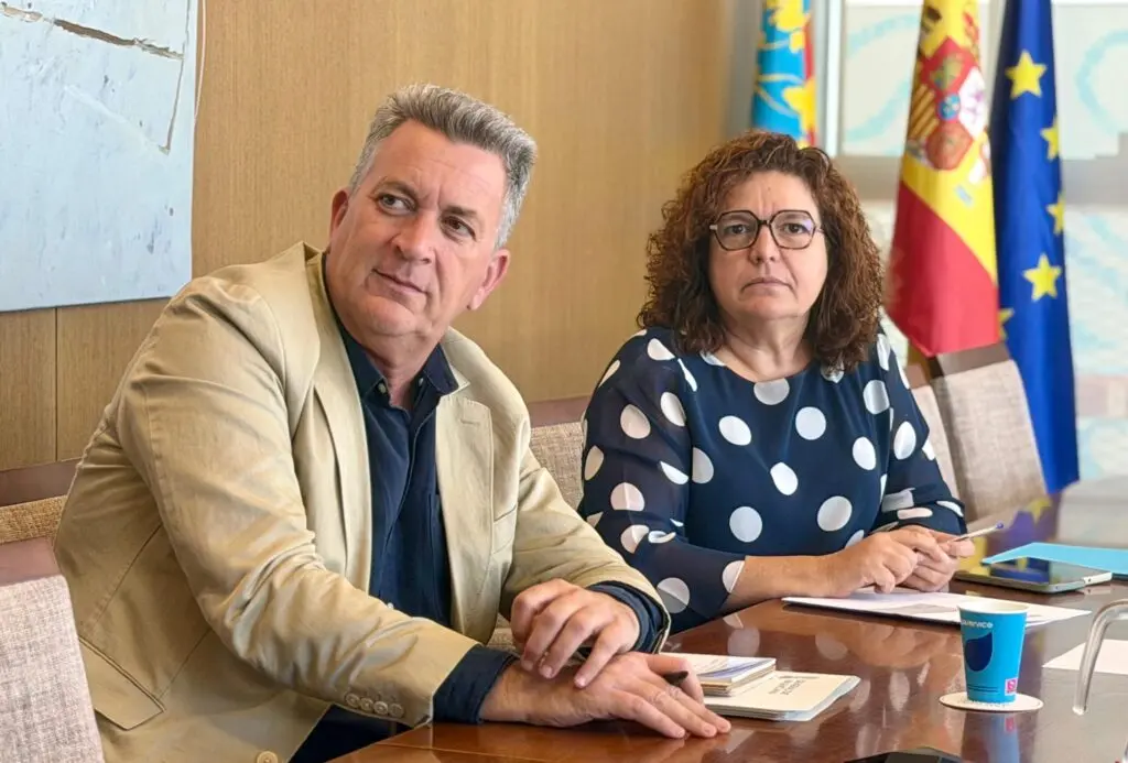 Dos personas sentadas a una mesa, con documentos y banderas al fondo.