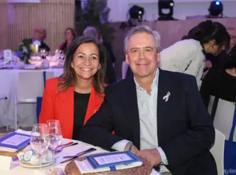 Dos personas sentadas en una mesa Dos personas posan sentadas ante una mesa puesta, con asistentes al fondo.