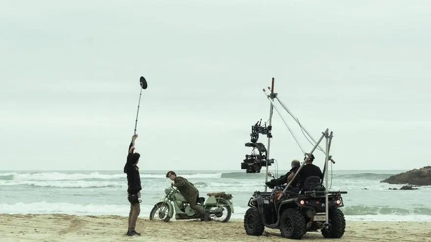 Equipo de rodaje con cámara y micrófono junto a una motocicleta en la playa.