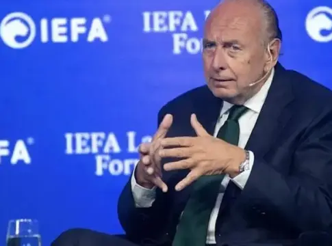 Hombre sentado ante un fondo de IEFA Hombre sentado en un escenario, con las manos juntas ante un fondo azul de IEFA.