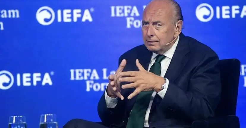 Hombre sentado ante un fondo de IEFA Hombre sentado en un escenario, con las manos juntas ante un fondo azul de IEFA.