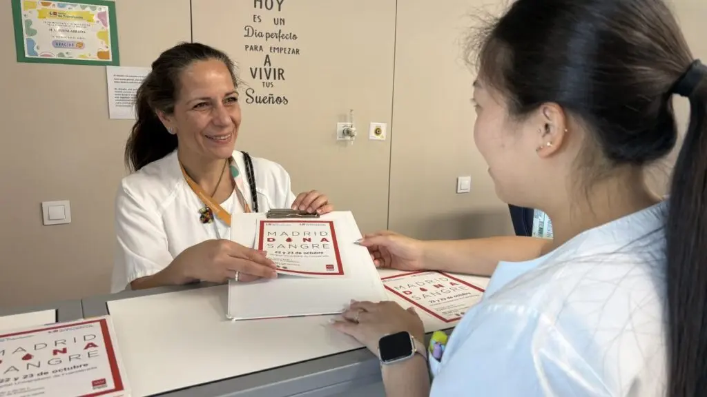 Dos mujeres intercambian un documento en un mostrador con carteles de donación de sangre.