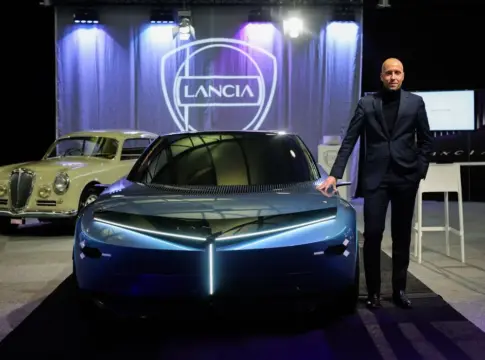 Automóviles Lancia en una exposición Hombre junto a un automóvil azul moderno, con un coche clásico y el logotipo Lancia detrás.