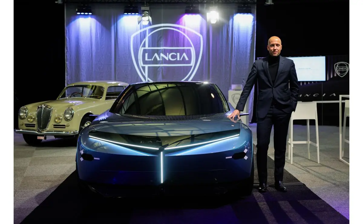 Hombre junto a un automóvil azul moderno, con un coche clásico y el logotipo Lancia detrás.