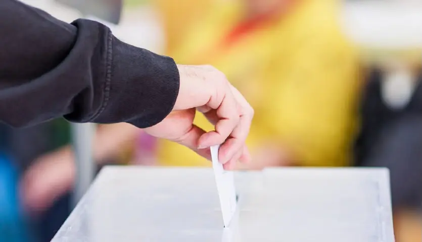 Una mano introduce una papeleta en una urna transparente durante una votación.