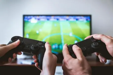 Dos mandos de videojuegos sostenidos frente a un televisor con un campo verde en pantalla