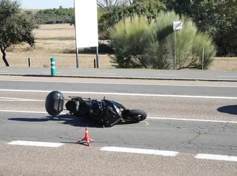Motocicleta caída sobre la calzada Una motocicleta negra yace de lado sobre la calzada, junto a un cono naranja.