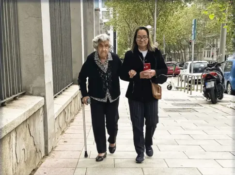 Dos mujeres caminan juntas por una acera Una mujer mayor camina con bastón junto a otra mujer por una acera urbana.