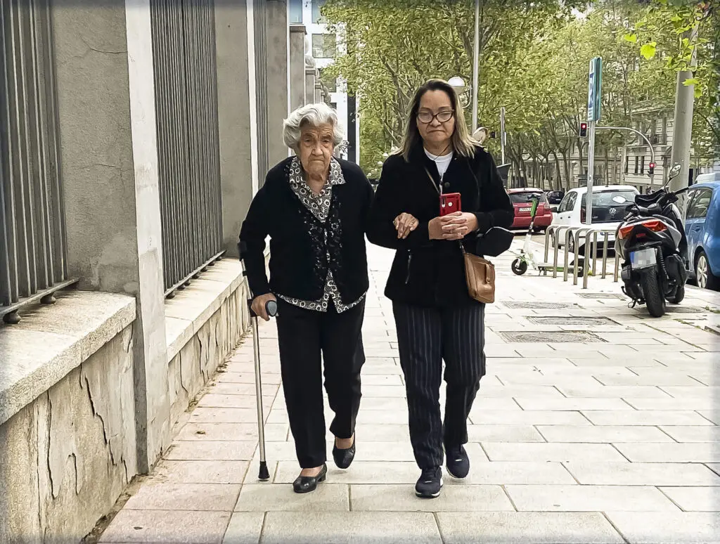 Una mujer mayor camina con bastón junto a otra mujer por una acera urbana.