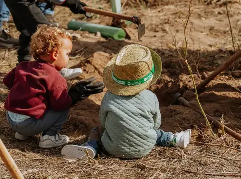 Niños junto a una actividad de plantación Dos niños observan una pequeña planta junto a tierra removida y herramientas de jardinería.