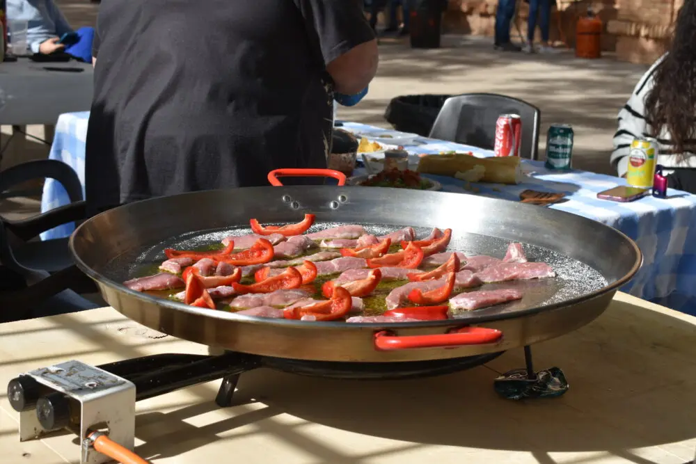 Paellera con ingredientes al aire libre Paellera grande con carne y pimiento rojo sobre un quemador al aire libre