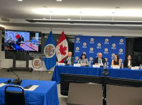 Panel en una sala con emblemas de la OEA Cinco personas sentadas en una mesa ante un panel azul con emblemas de la OEA.