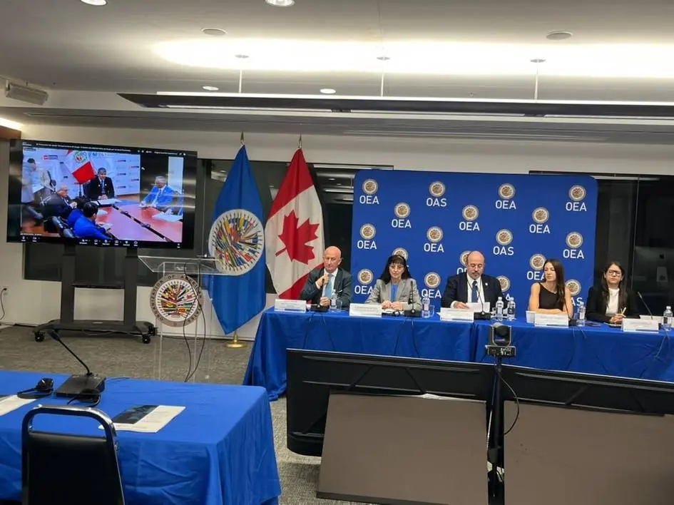 Panel en una sala con emblemas de la OEA Cinco personas sentadas en una mesa ante un panel azul con emblemas de la OEA.