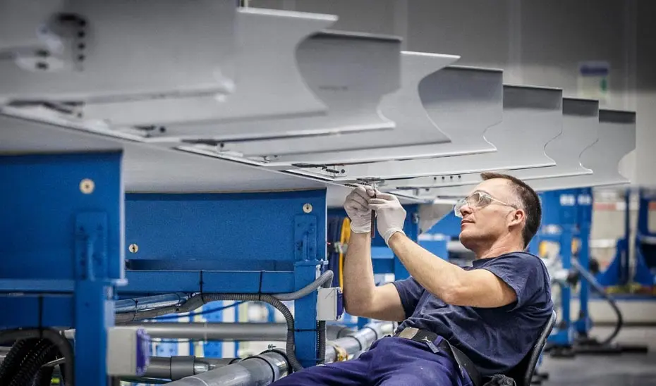 Una persona con gafas y guantes manipula una pieza bajo maquinaria industrial.