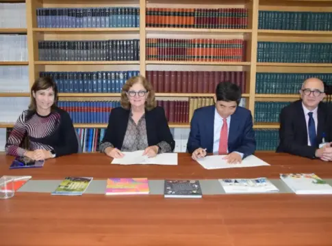 Personas escriben documentos ante estanterías Cuatro personas se sientan ante una mesa; dos escriben en documentos.