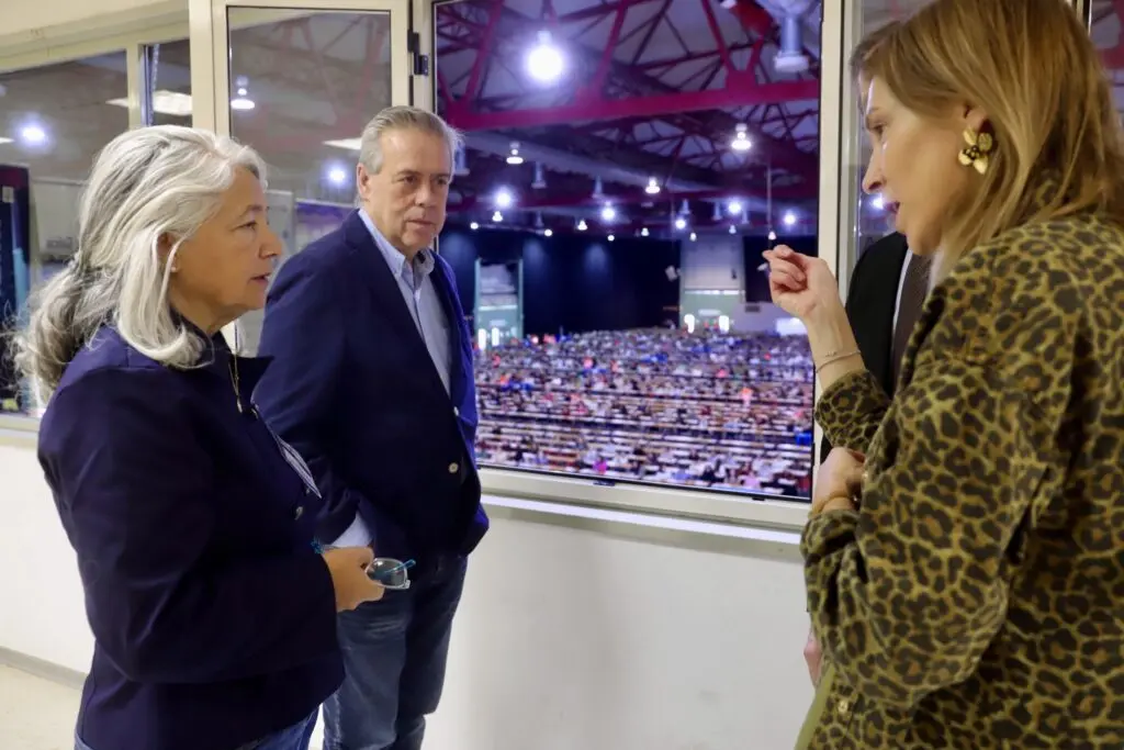 Tres personas conversan junto a un ventanal con una sala llena de mesas al fondo.