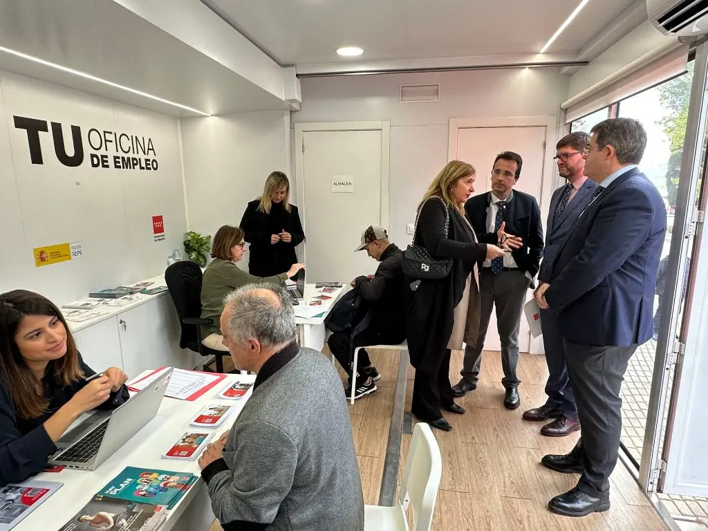 Personas conversan junto a mesas con folletos en una oficina rotulada de empleo.