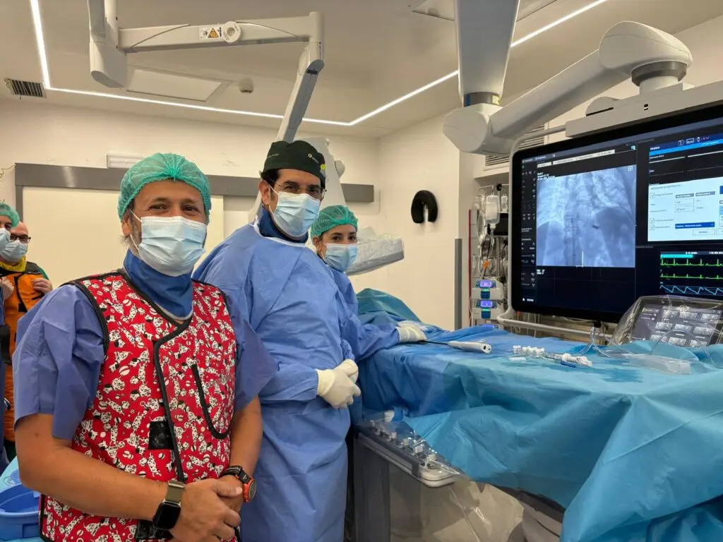 Personas en una sala de procedimientos Personas con mascarillas junto a una mesa cubierta y un monitor médico