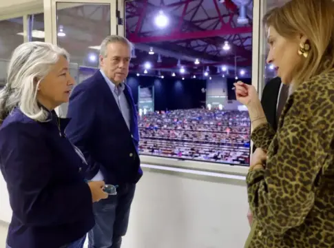 Personas observan una sala con filas de mesas Tres personas observan desde una ventana una sala amplia con filas de mesas y asistentes.