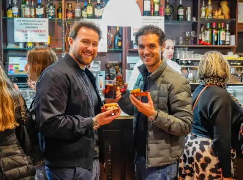 Personas con tapas en una barra Dos hombres sostienen pequeñas tapas y bebidas frente a una barra de bar.