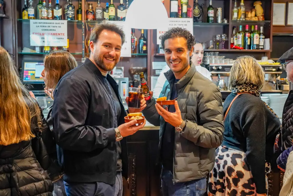 Dos hombres sostienen pequeñas tapas y bebidas frente a una barra de bar.