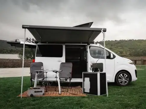 Furgoneta camper blanca con toldo y mobiliario exterior Furgoneta camper blanca con toldo desplegado, mesa y sillas en una zona verde.