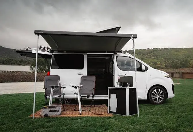 Furgoneta camper blanca con toldo y mobiliario exterior Furgoneta camper blanca con toldo desplegado, mesa y sillas en una zona verde.