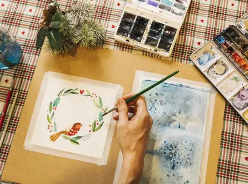 Pintura navideña con acuarelas Una mano pinta una ilustración navideña con acuarelas sobre una mesa de cuadros.