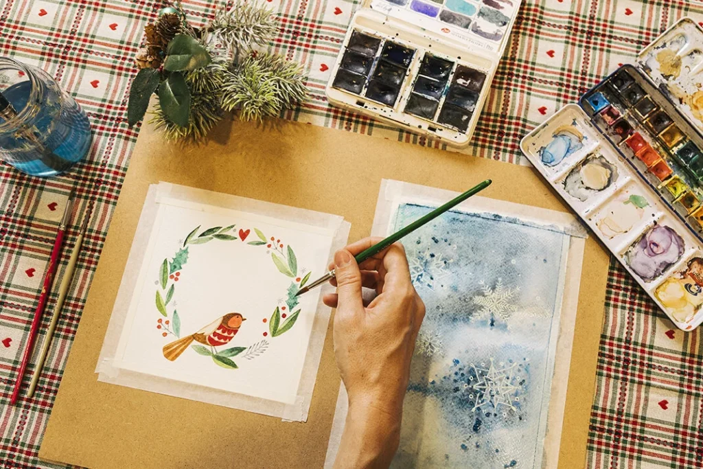 Pintura navideña con acuarelas Una mano pinta una ilustración navideña con acuarelas sobre una mesa de cuadros.