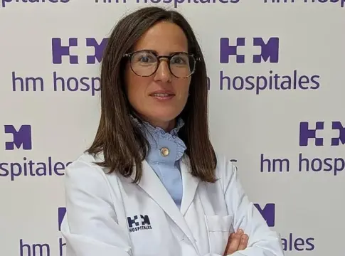 Retrato de Teresa Hernández ante HM Hospitales Mujer con bata blanca y gafas posa ante un panel de HM Hospitales.