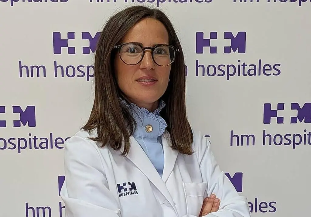Mujer con bata blanca y gafas posa ante un panel de HM Hospitales.