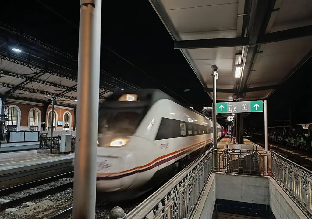 Un tren blanco pasa por una estación ferroviaria iluminada durante la noche.