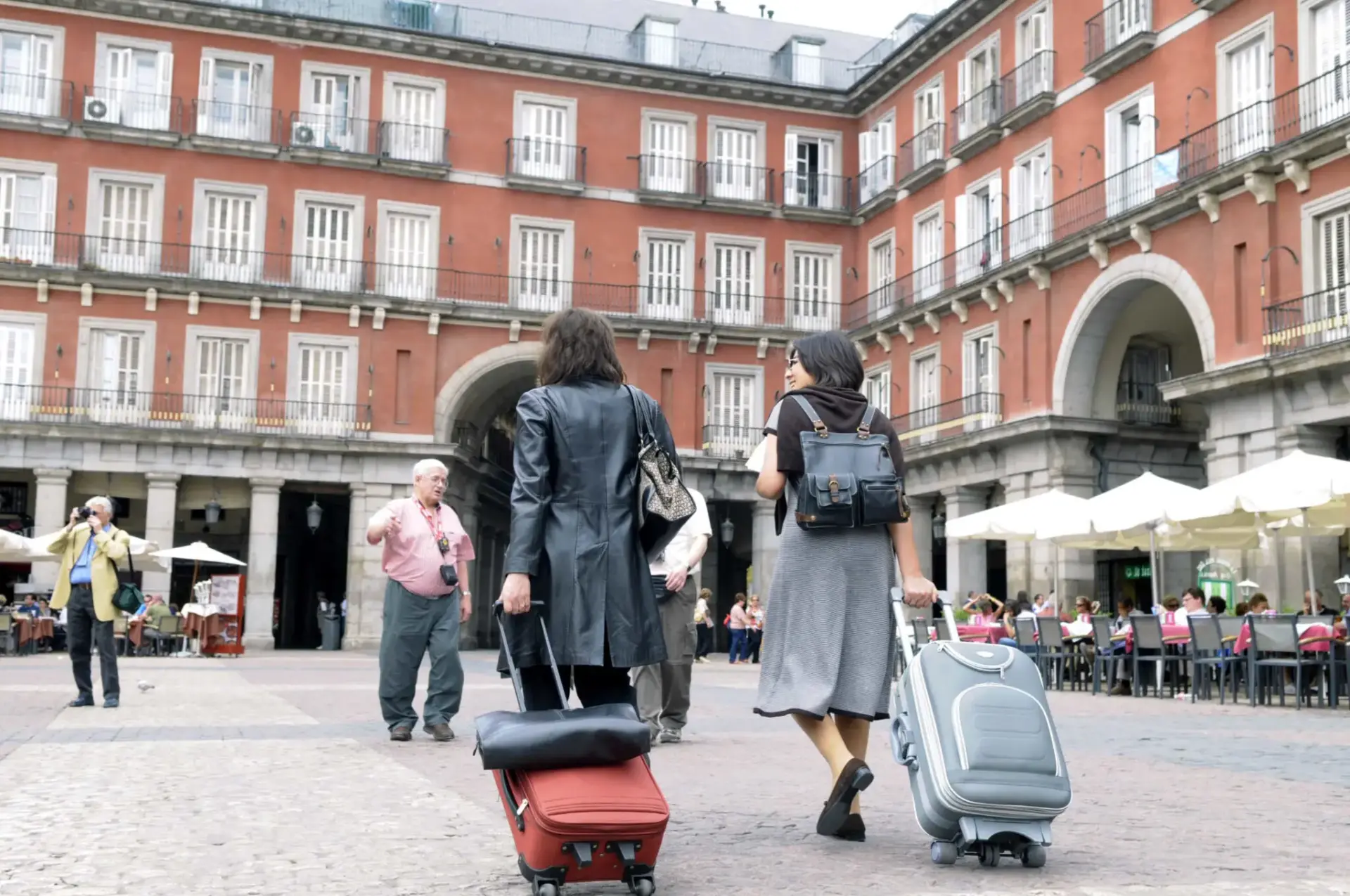 Viajeros con maletas en la Plaza Mayor Dos personas con maletas de ruedas cruzan la Plaza Mayor, rodeadas de edificios con balcones.