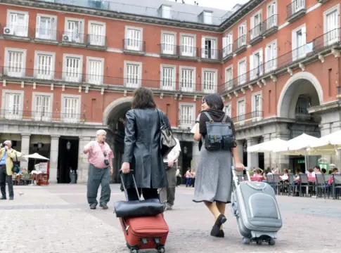 Viajeros con maletas en la Plaza Mayor Dos personas con maletas de ruedas cruzan la Plaza Mayor, rodeadas de edificios con balcones.