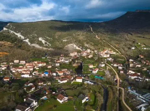 Pueblo rural entre campos y montañas Vista aérea de un pequeño pueblo entre campos y laderas boscosas bajo un cielo nublado.